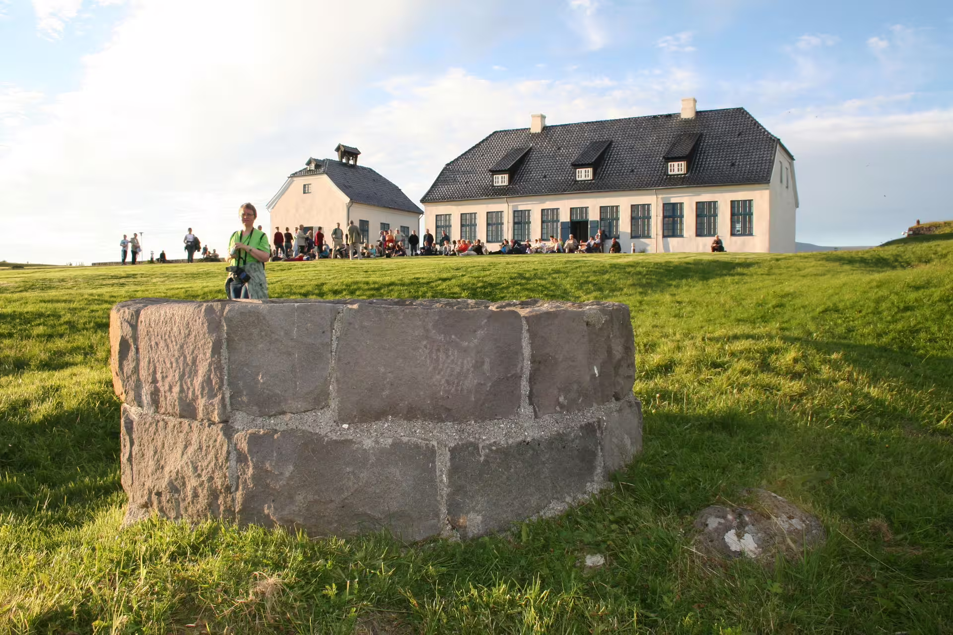 Stone well on Videy Island with historic white building and visitors in background, Reykjavik