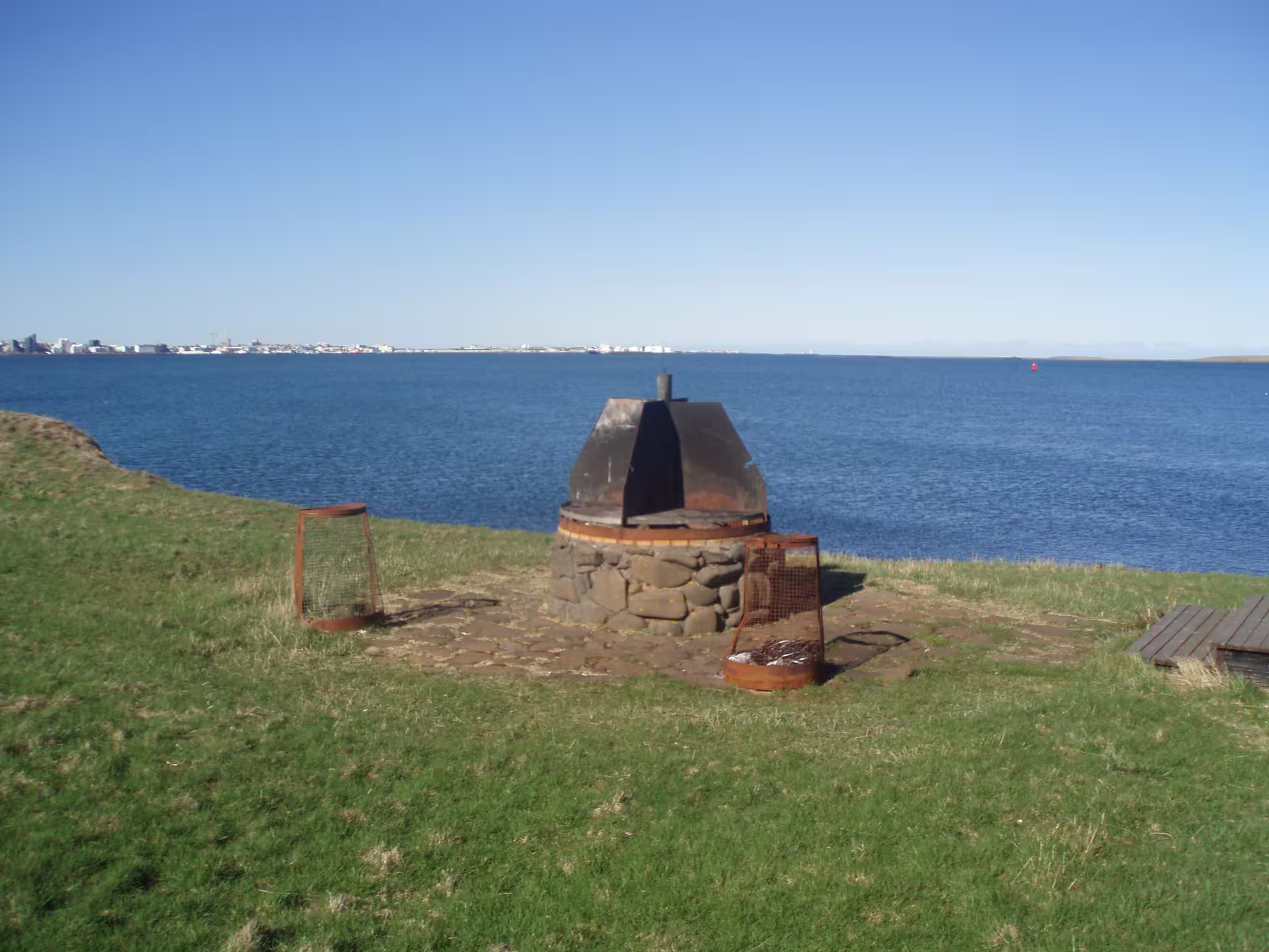 Stone firepit with metal hood on Videy Island grassy shoreline overlooking Reykjavik across the bay