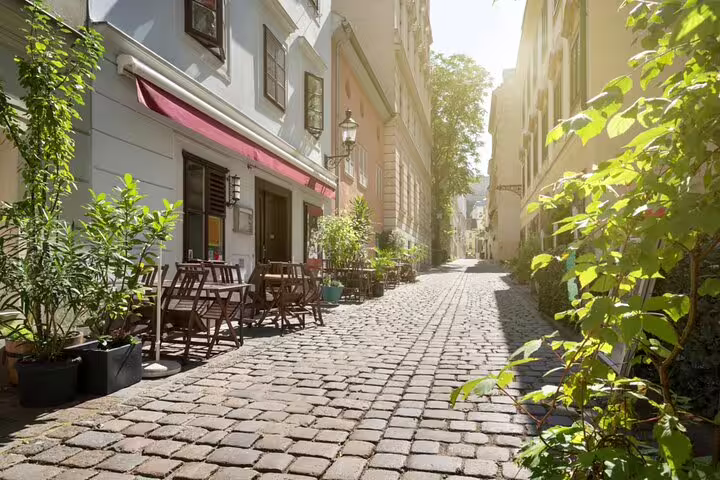 Sunlit cobblestone alley in Vienna with outdoor café seating and green plants along historic buildings