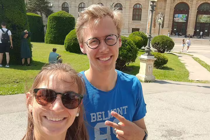 Two smiling tourists taking a selfie during a walking tour near a grand Vienna museum building