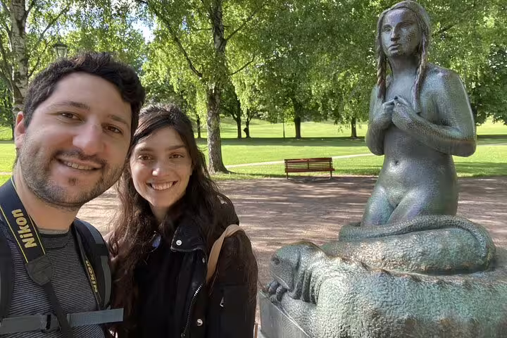 Couple taking selfie with bronze mermaid statue in Vigeland Park, Oslo, surrounded by green trees