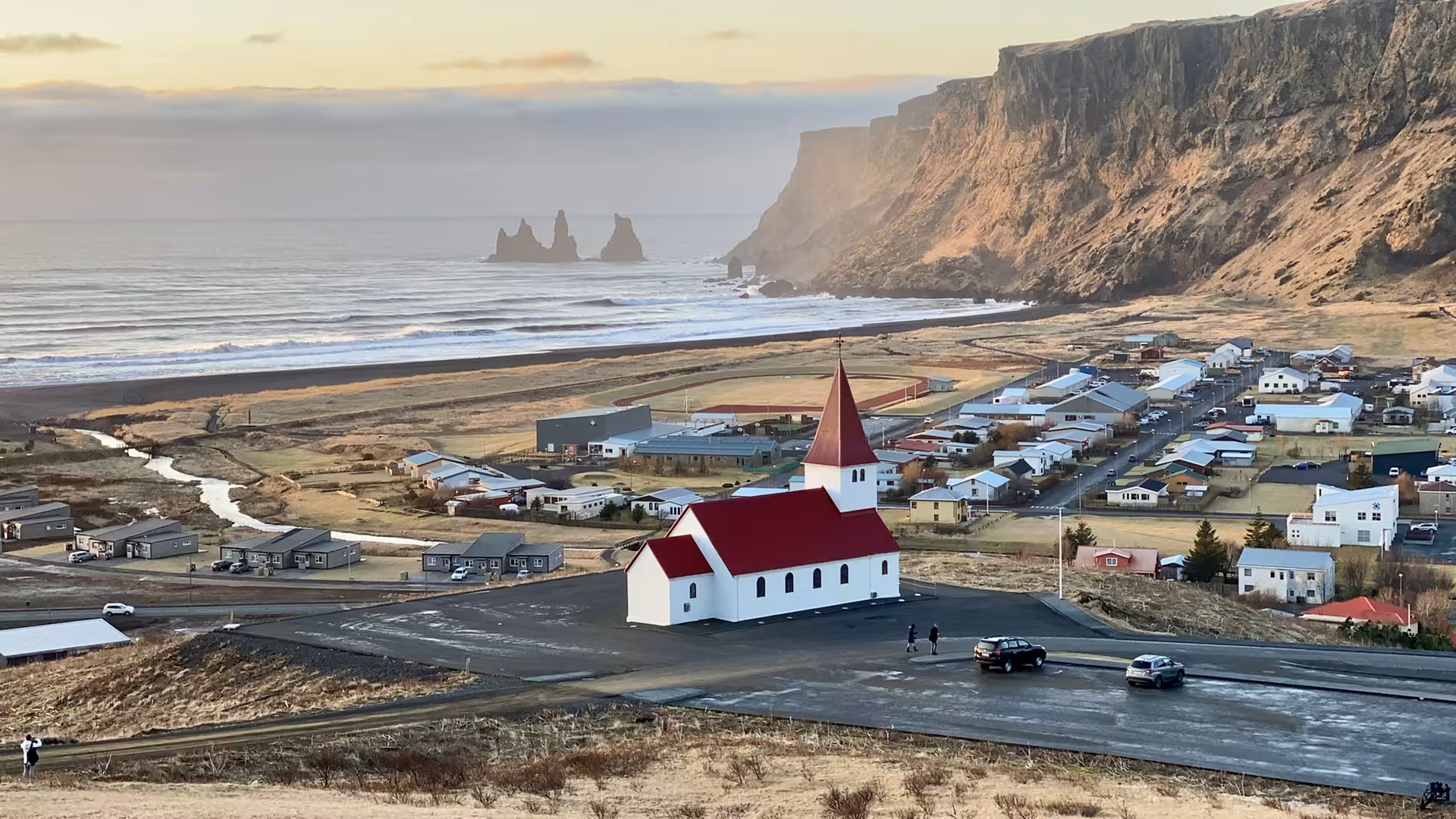 Aerial view of Vík í Mýrdal church with red roof, black sand beach, and Reynisdrangar sea stacks, Iceland South Coast