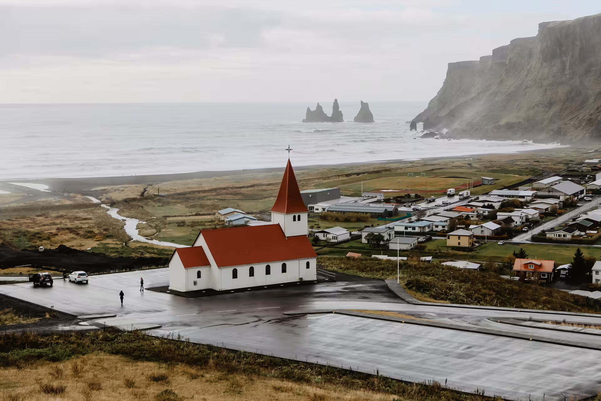 Aerial view of Vík í Mýrdal church with red roof, black sand beach, and Reynisdrangar sea stacks, Iceland