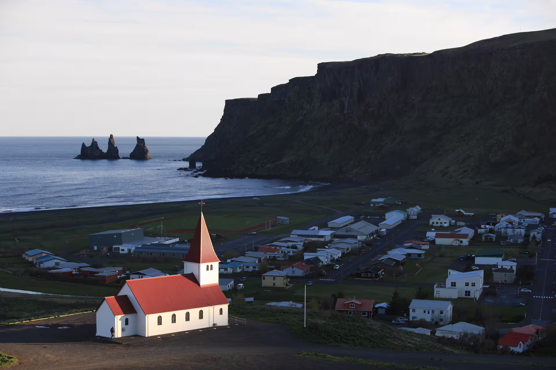 Aerial view of Vík village with red-roofed church, black sand beach, Reynisdrangar sea stacks, and dramatic cliffs in South Iceland