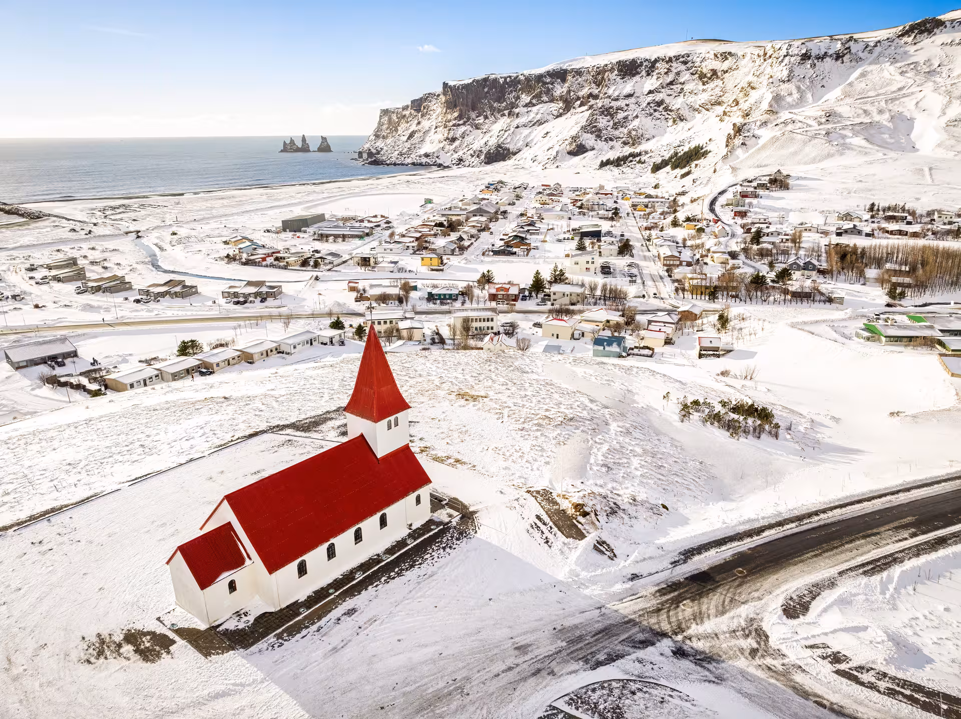 Aerial view of Vík í Mýrdal village with red-roofed church, black sand beach, and sea stacks in winter, Iceland