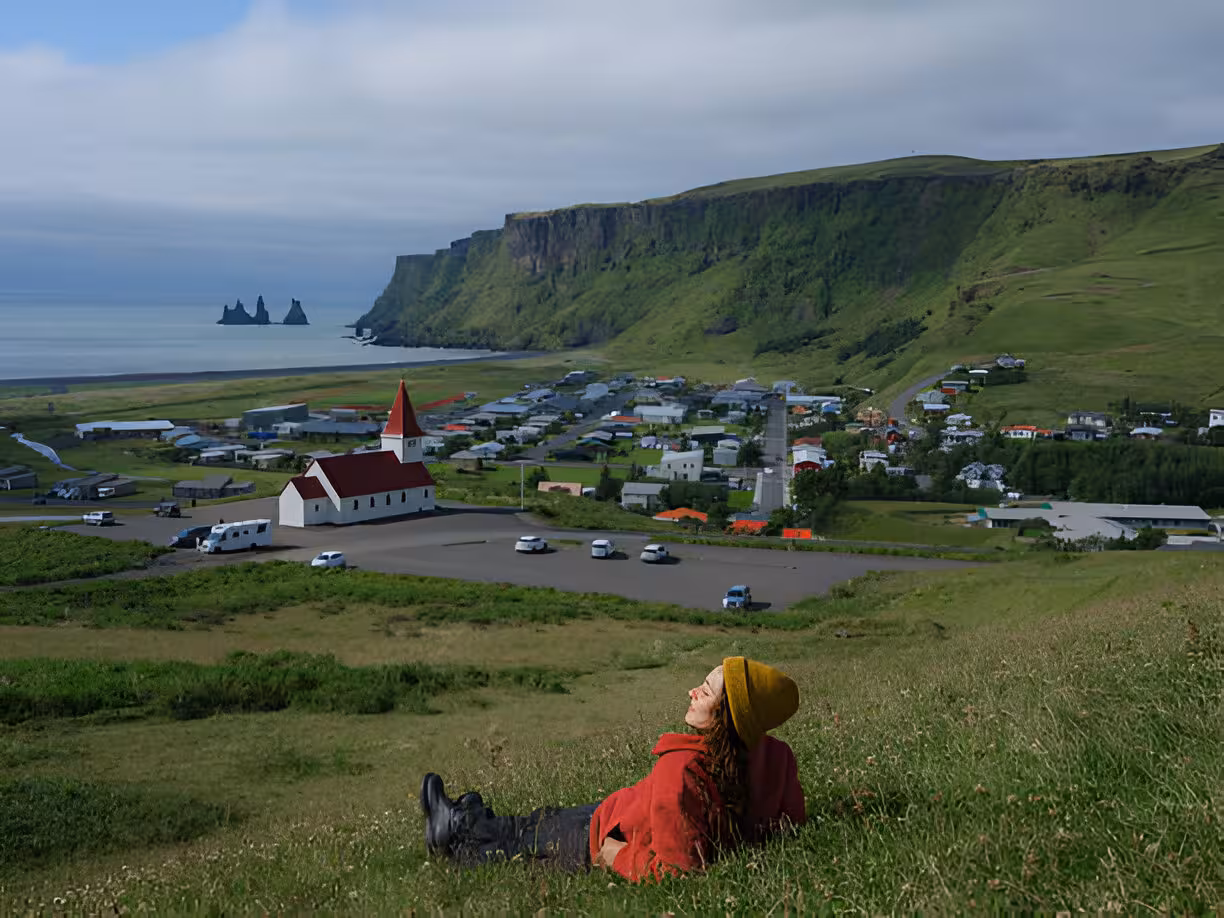 Traveler resting on hillside above Vík village with black sand beach and sea stacks in Iceland