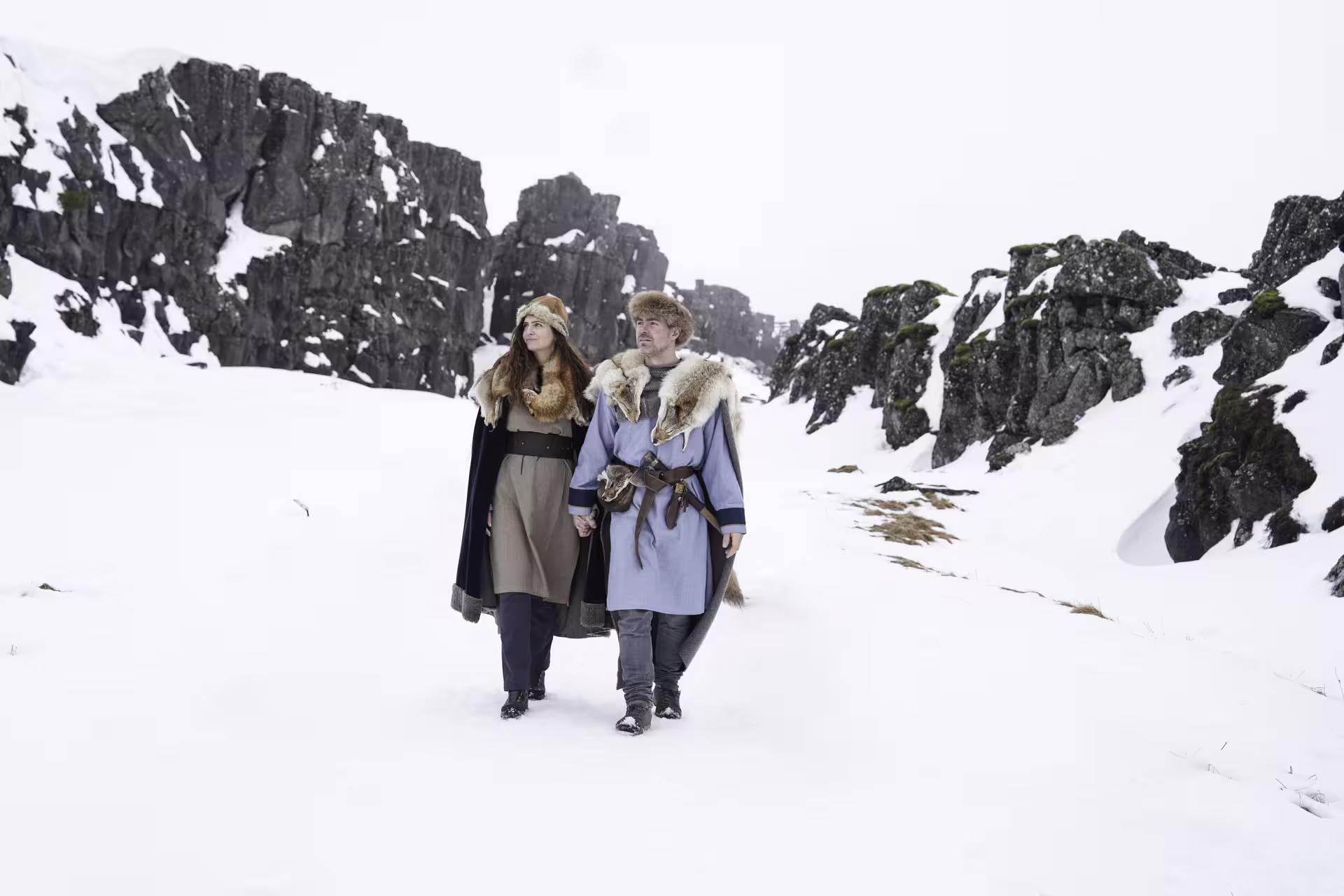 Couple dressed in Viking fur costumes walking through snowy Iceland landscape with dramatic rocky cliffs on circle tour
