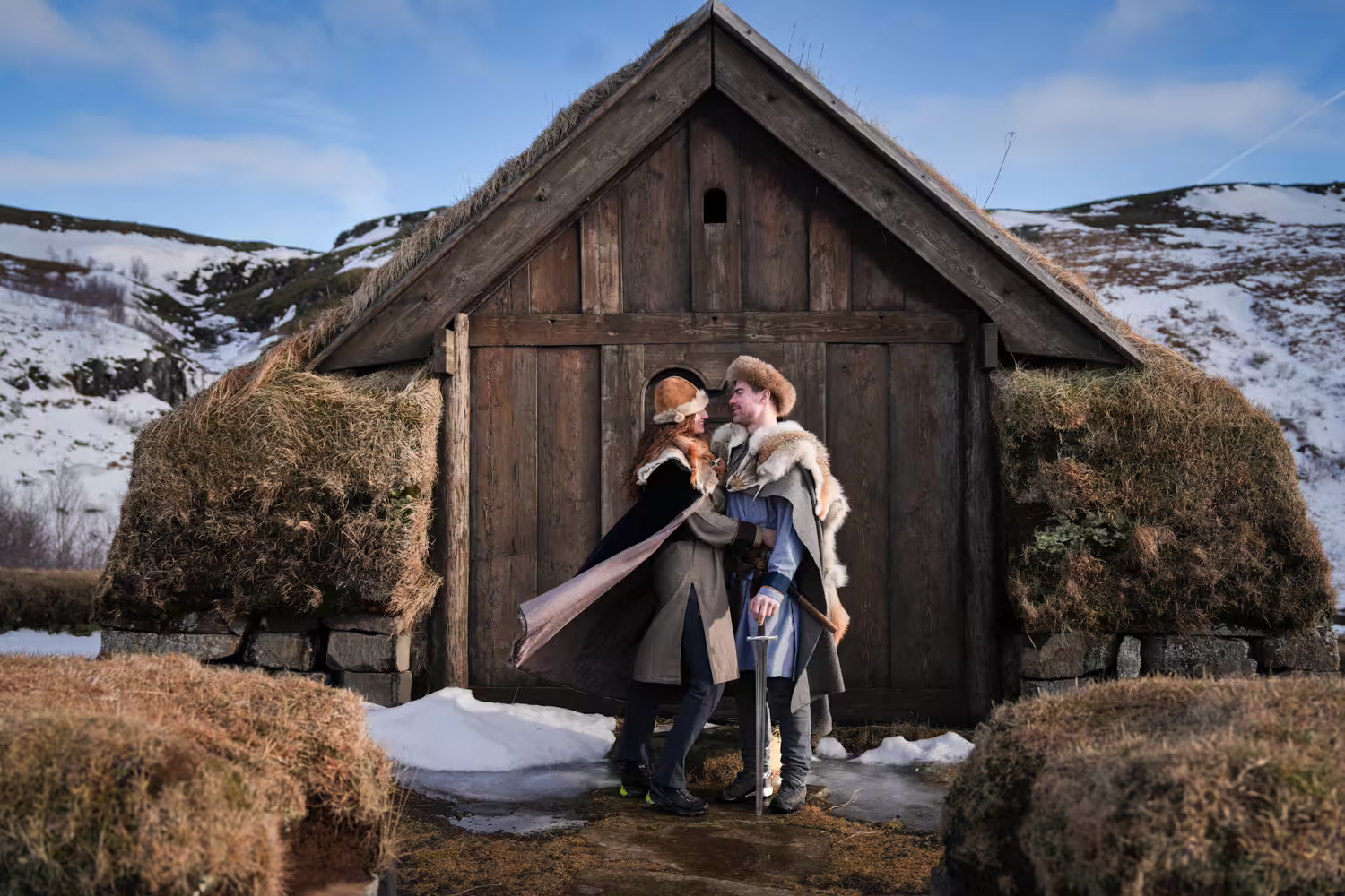 Couple in Viking costumes posing before a historic turf-roofed Norse longhouse on a snowy Celtic journey