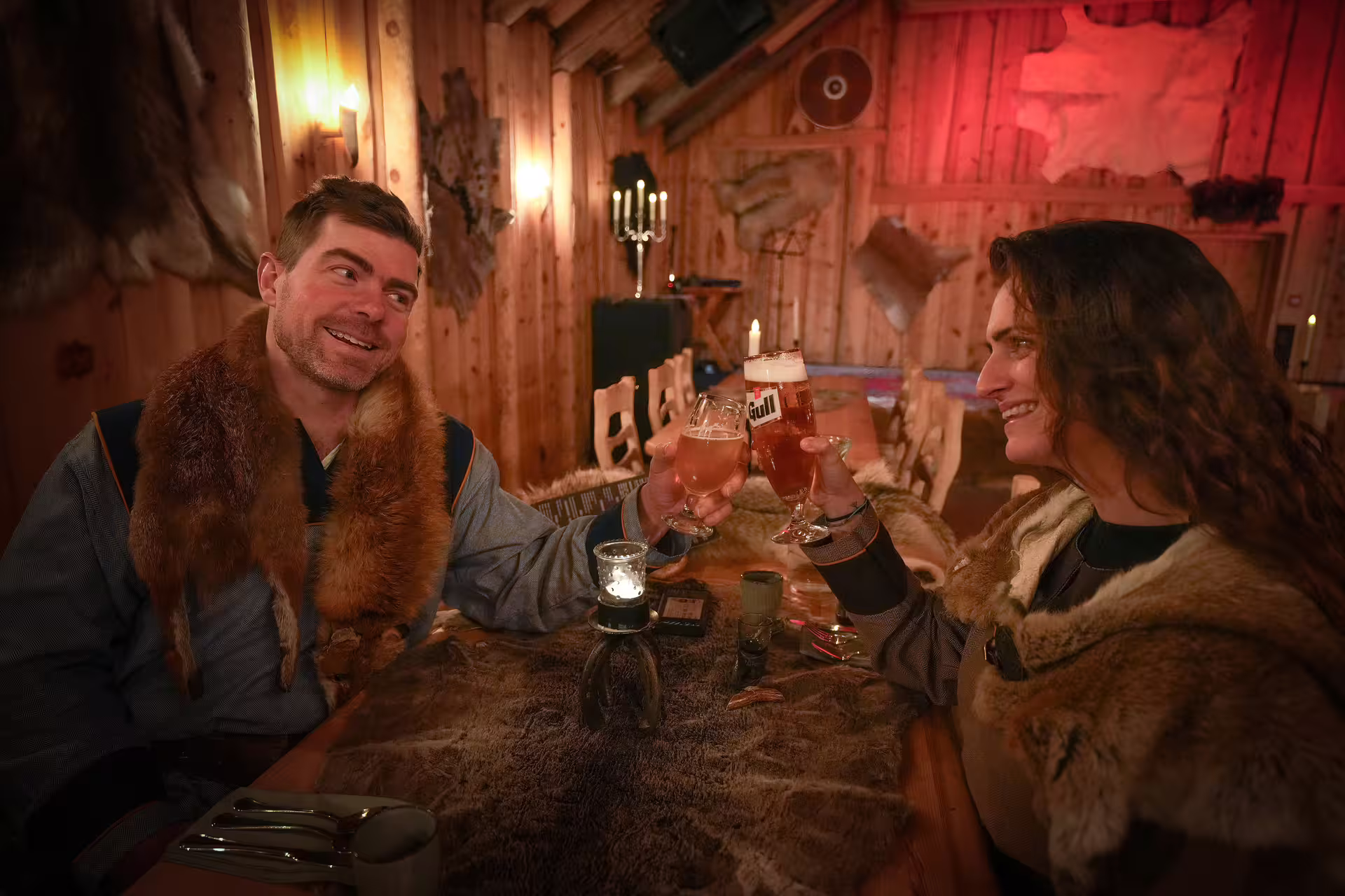 Couple in Viking fur costumes toasting drinks at a rustic Norse longhouse during Iceland Viking circle tour experience