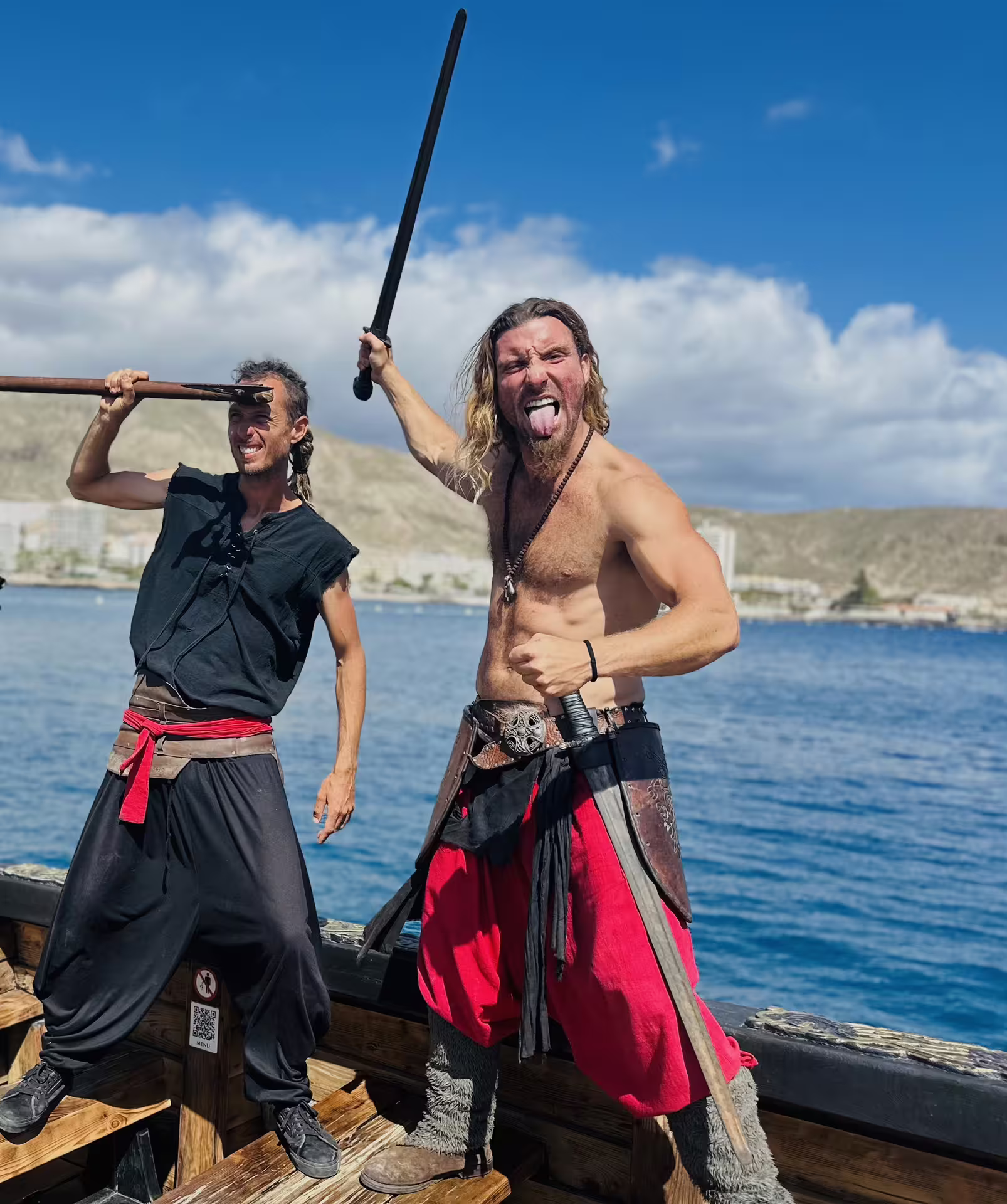 Two enthusiastic Viking actors with swords pose playfully on a ship with scenic ocean views.