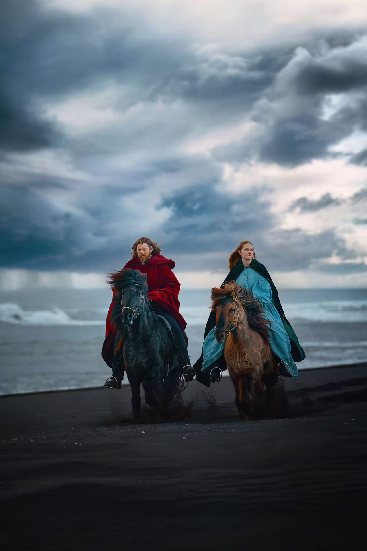 Two riders in Viking cloaks on Icelandic horses galloping along dramatic black sand beach South Coast