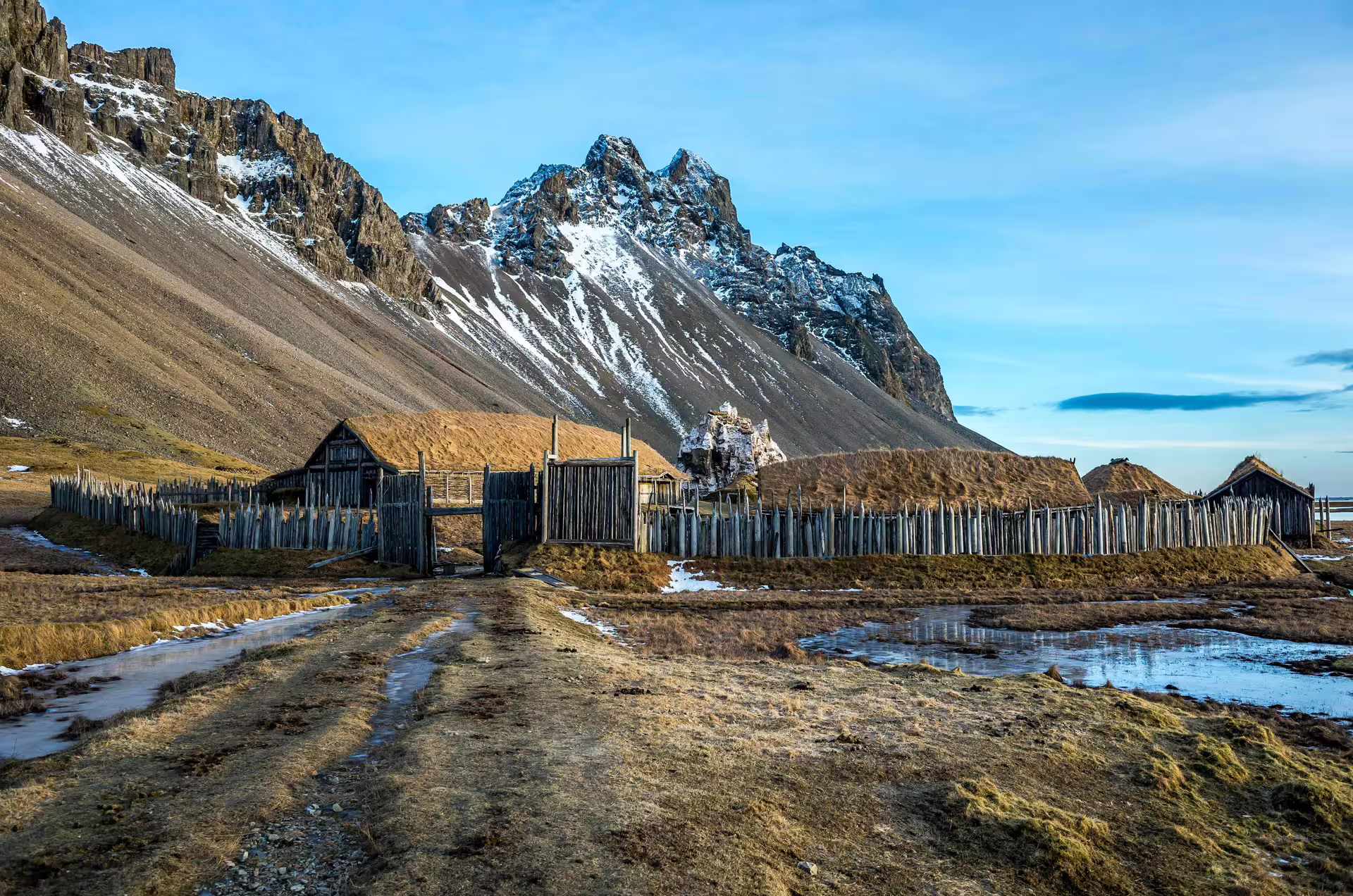 Historic Viking village with turf-roofed longhouses and wooden palisade beneath dramatic snow-capped Vestrahorn mountain in Southeast Iceland