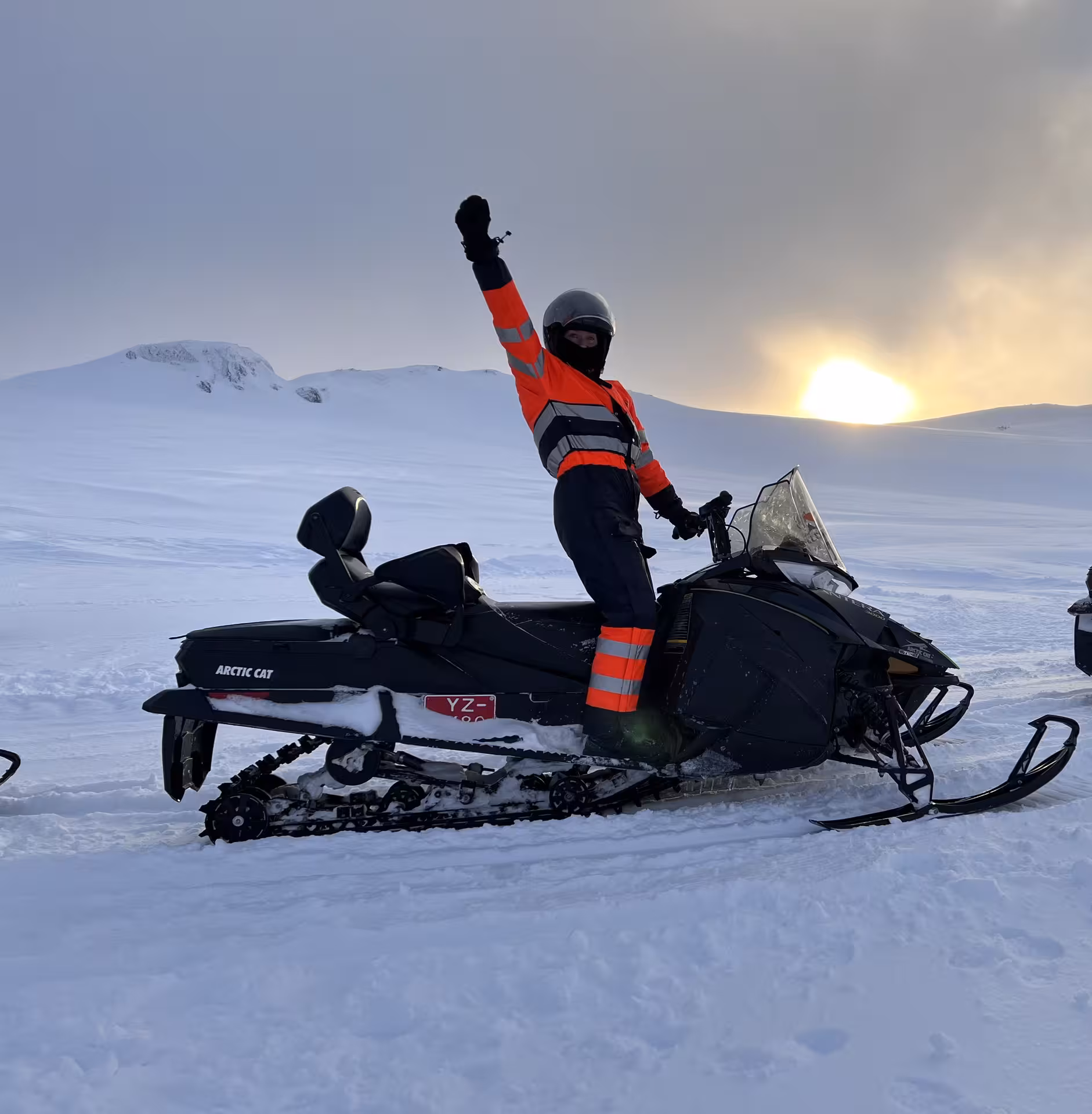 Woman striking a victorious pose on a snowmobile against a stunning snowy landscape at sunset in Iceland.