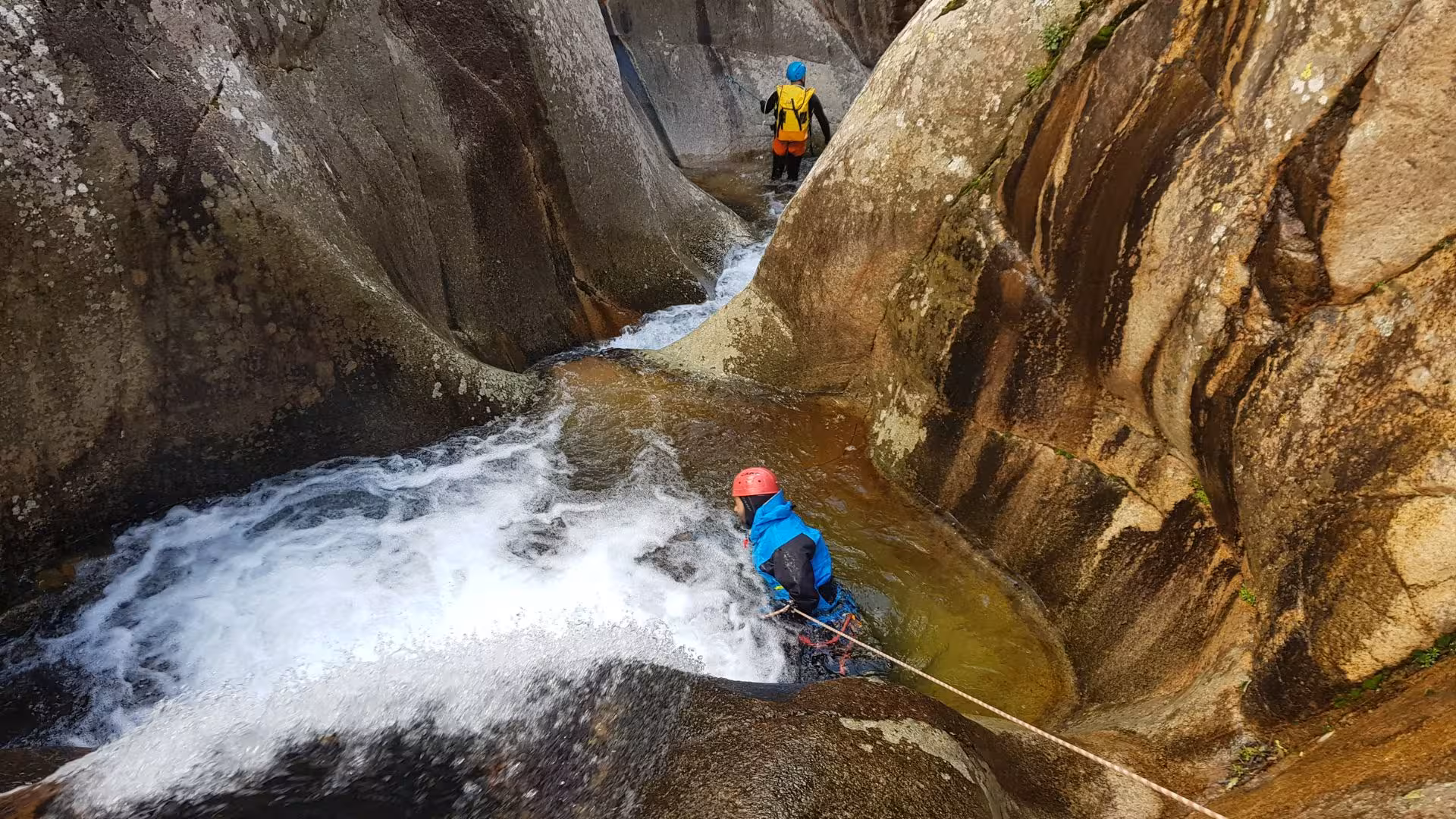 Two participants descending a narrow water chute in Villacidro's Rio Zairi canyoning experience, showcasing natural beauty.