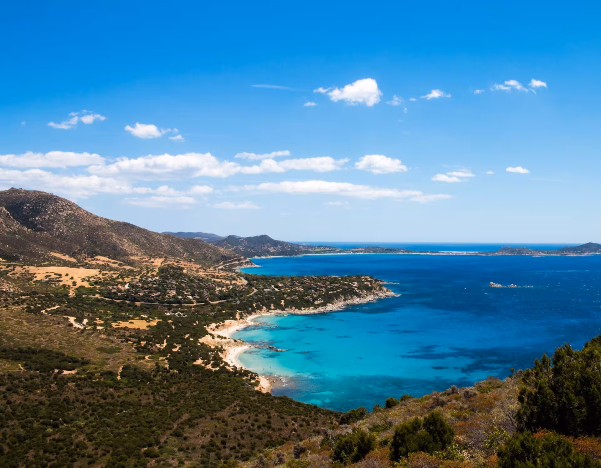 Panoramic view of Villasimius coastline with azure waters and rolling hills under a bright blue sky in Sardinia, Italy.