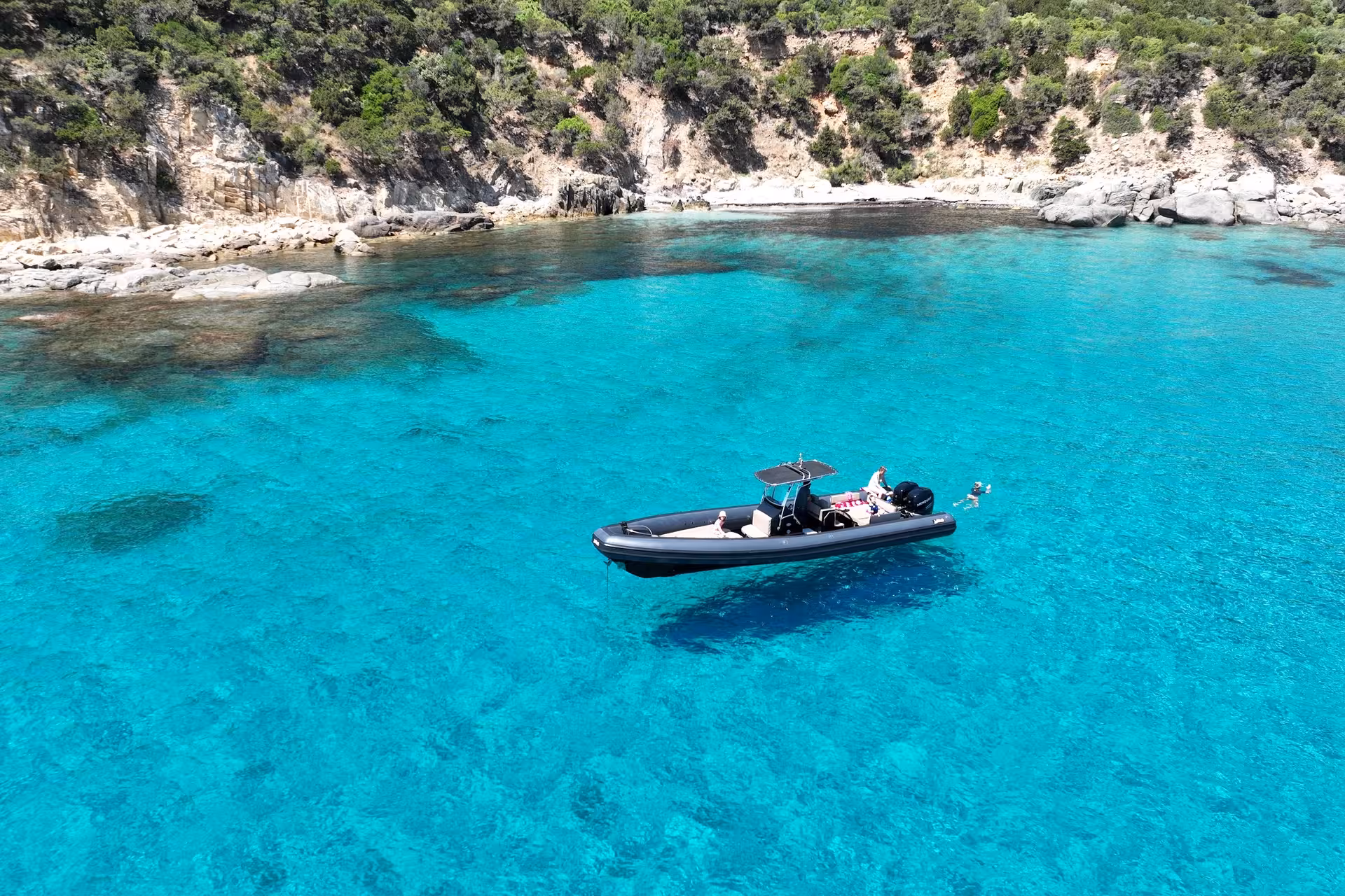 RIB anchored in crystal-clear waters with people snorkeling near rocky shores on Villasimius tour.