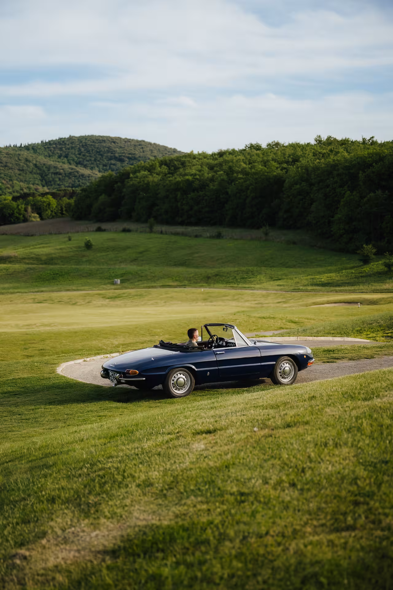 Navy blue vintage Alfa Romeo Spider convertible driving through green rolling hills countryside