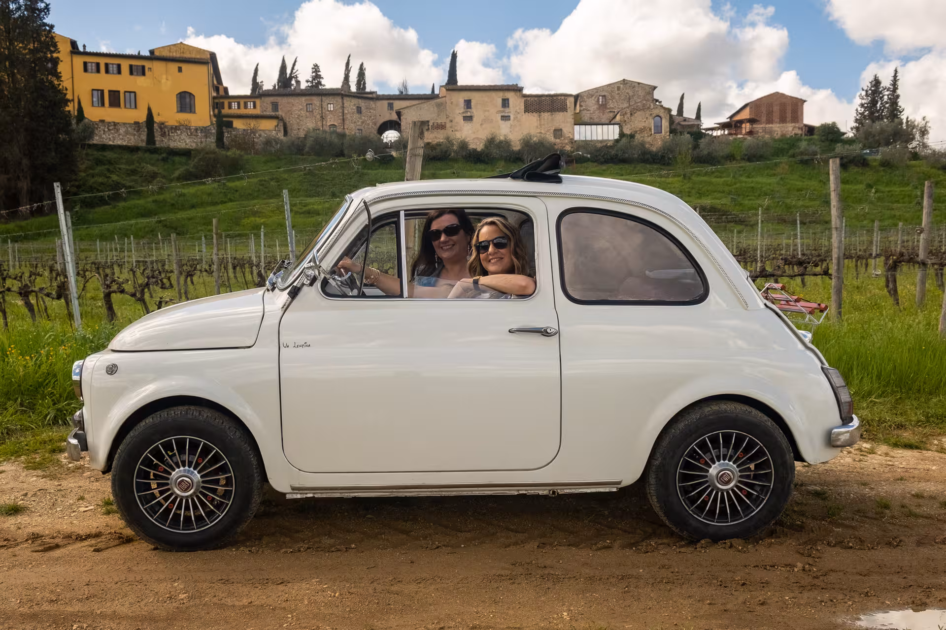 Two women smiling inside a vintage white Fiat 500 parked in a scenic Tuscan vineyard.
