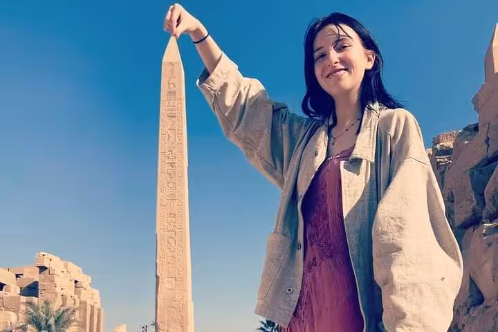 Smiling visitor playfully poses with the towering obelisk at Luxor's iconic Karnak Temple under a bright sky.