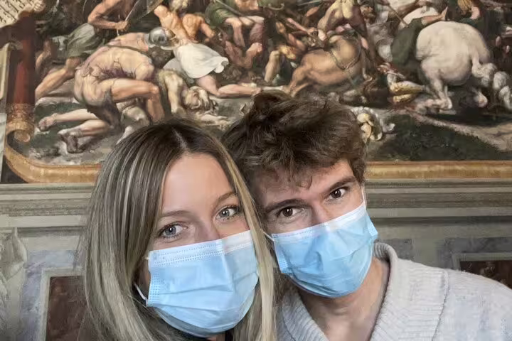 Two visitors wearing blue face masks taking a selfie inside the Vatican Museums with a Renaissance fresco mural behind them