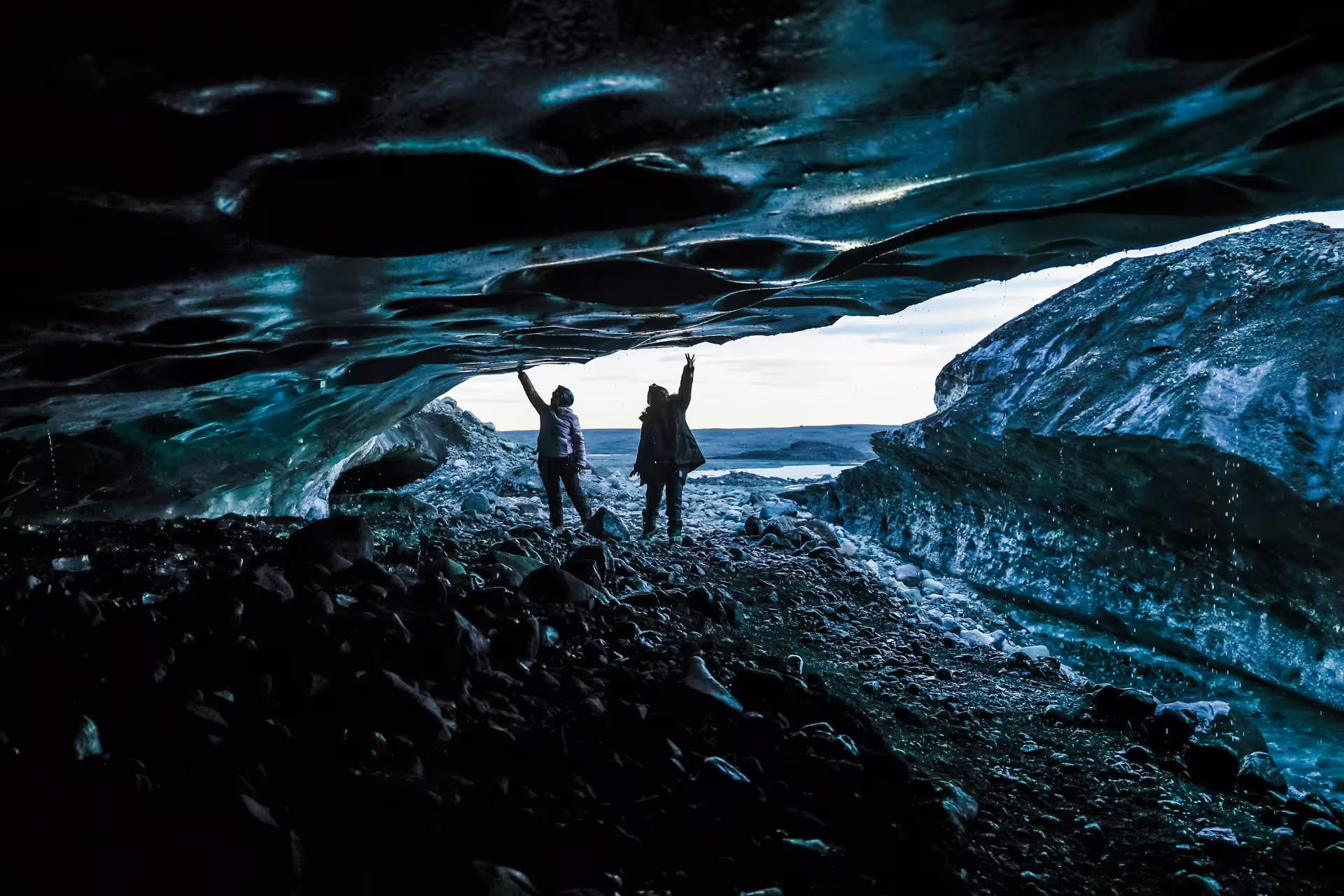 Two visitors silhouetted at the entrance of a blue glacial ice cave reaching up to touch the ice on a private tour