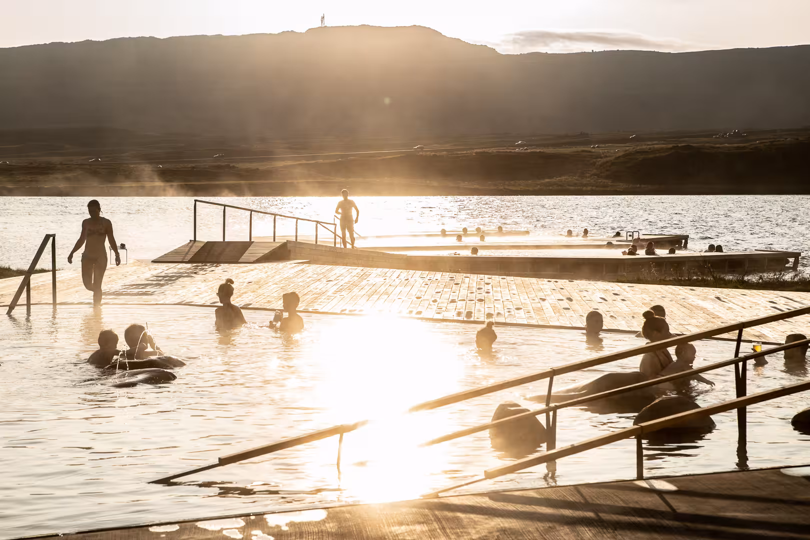 Visitors soaking in Vok Baths geothermal pool at golden sunset with lake and mountains in East Iceland