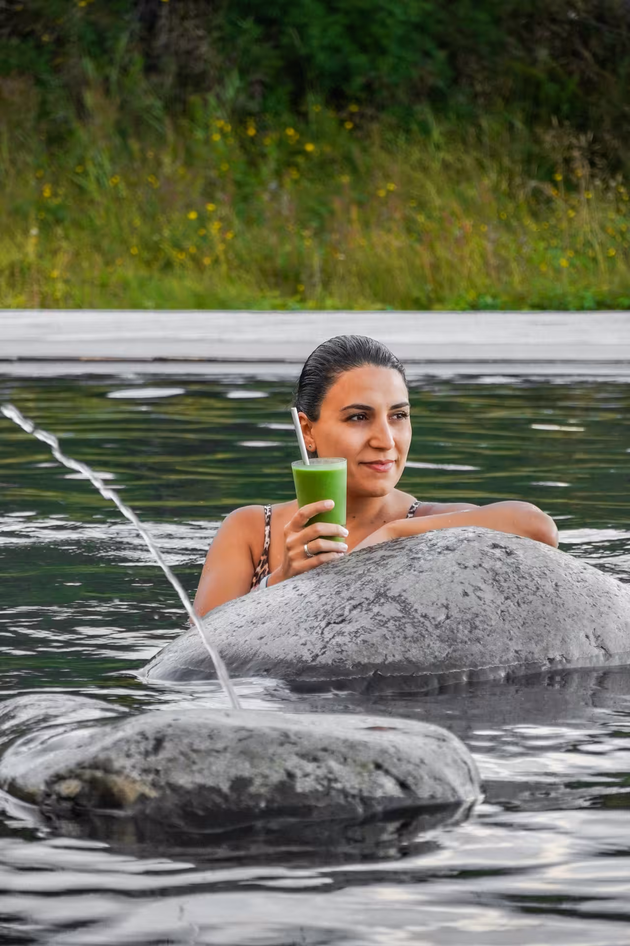 Woman relaxing in Vok Baths geothermal pool holding green smoothie surrounded by rocks