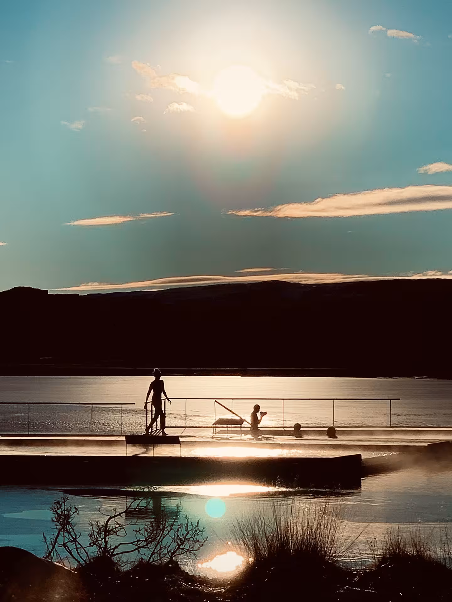 Silhouettes of people at Vok Baths geothermal pool overlooking a sunlit fjord in Iceland