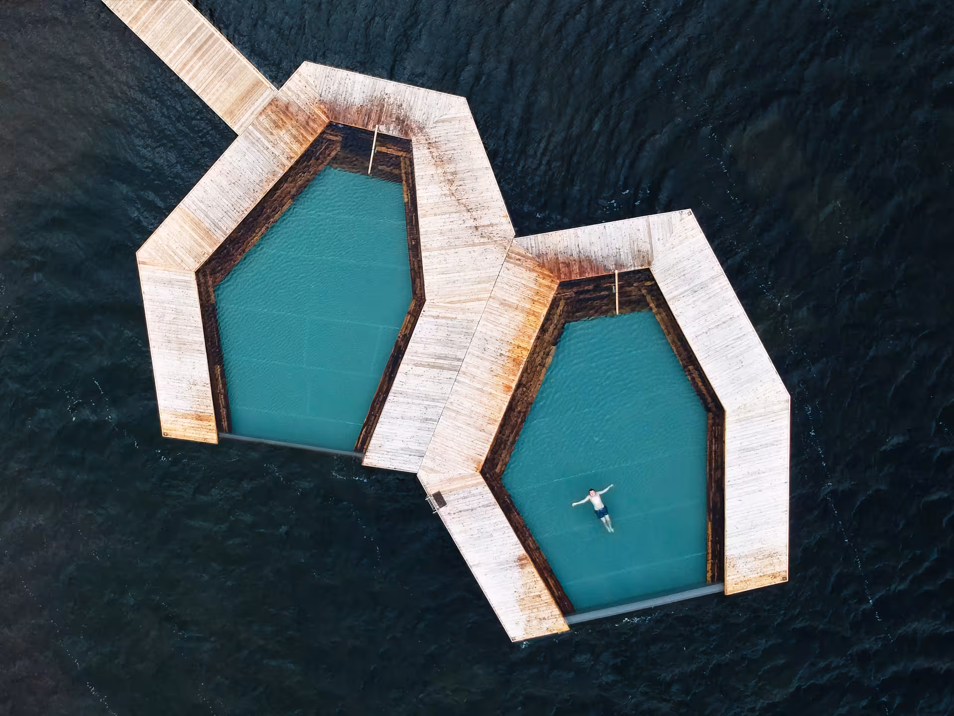 Aerial view of hexagonal Vok Baths floating pools with turquoise water and a swimmer in Eastfjords Iceland