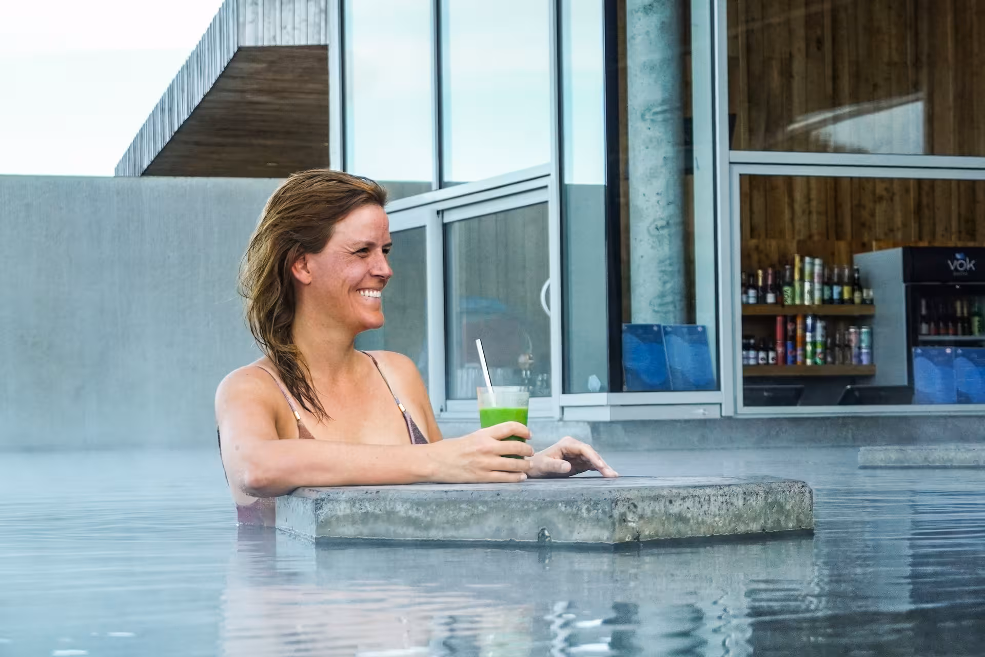Woman relaxing in Vok Baths geothermal pool holding green drink near poolside bar