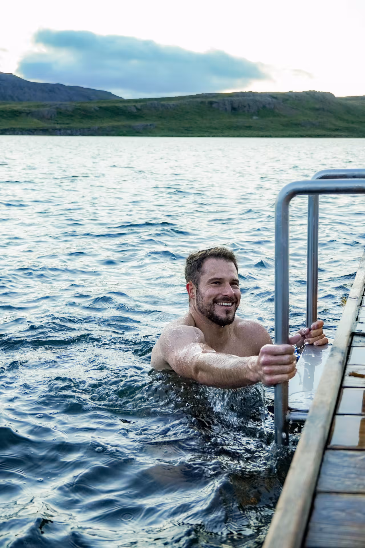 Smiling man swimming in Vok Baths lake with scenic East Iceland mountains in background