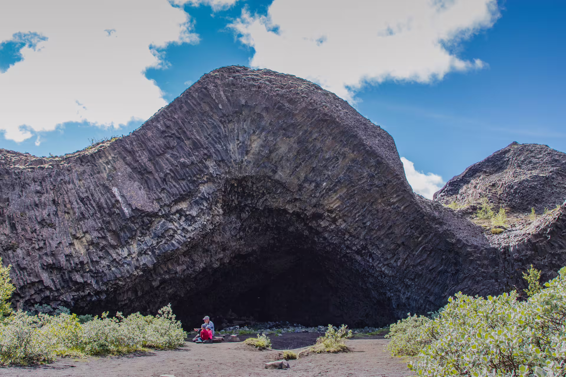 Two visitors sitting at the entrance of a massive volcanic basalt cave arch in North Iceland