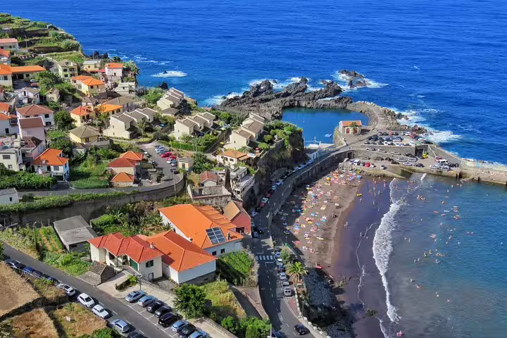 Aerial view of scenic coastal village with red-roofed houses, beachgoers, volcanic pools, and rugged cliffs by the Atlantic Ocean.