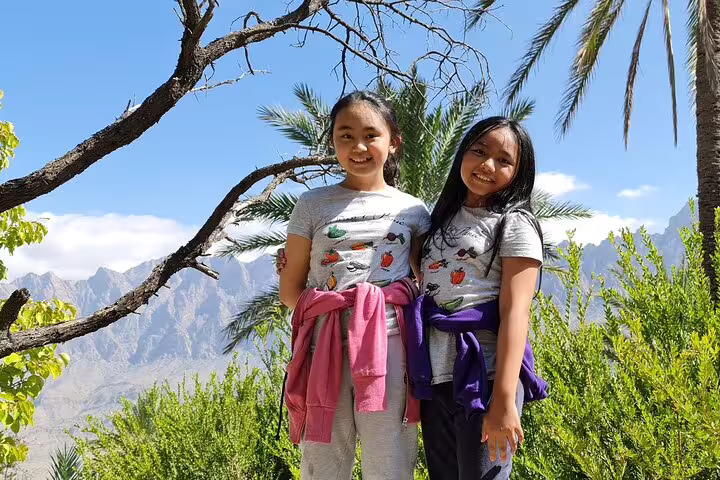 Two smiling children posing amidst lush greenery and palm trees at Wakan Village, with mountains in the background.