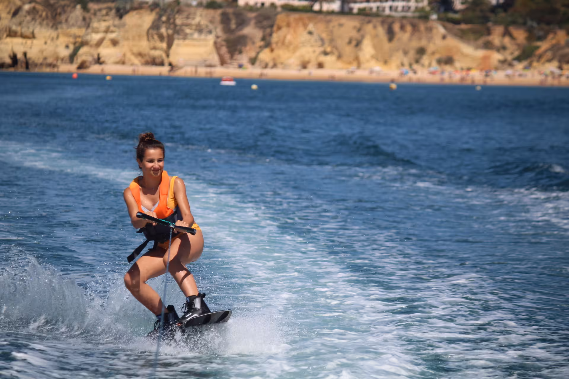 Smiling girl in orange life vest wakeboarding on sparkling blue waters near Armacao de Pera cliffs