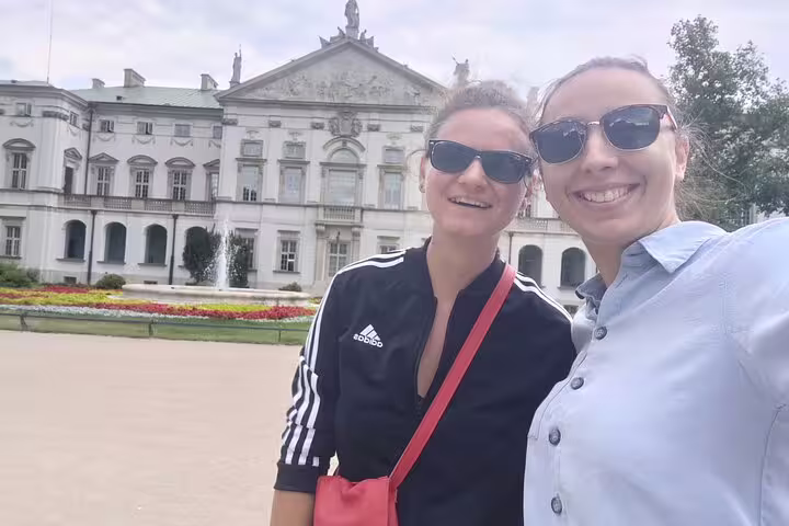 Two smiling women in sunglasses posing in front of a grand historic palace in Warsaw