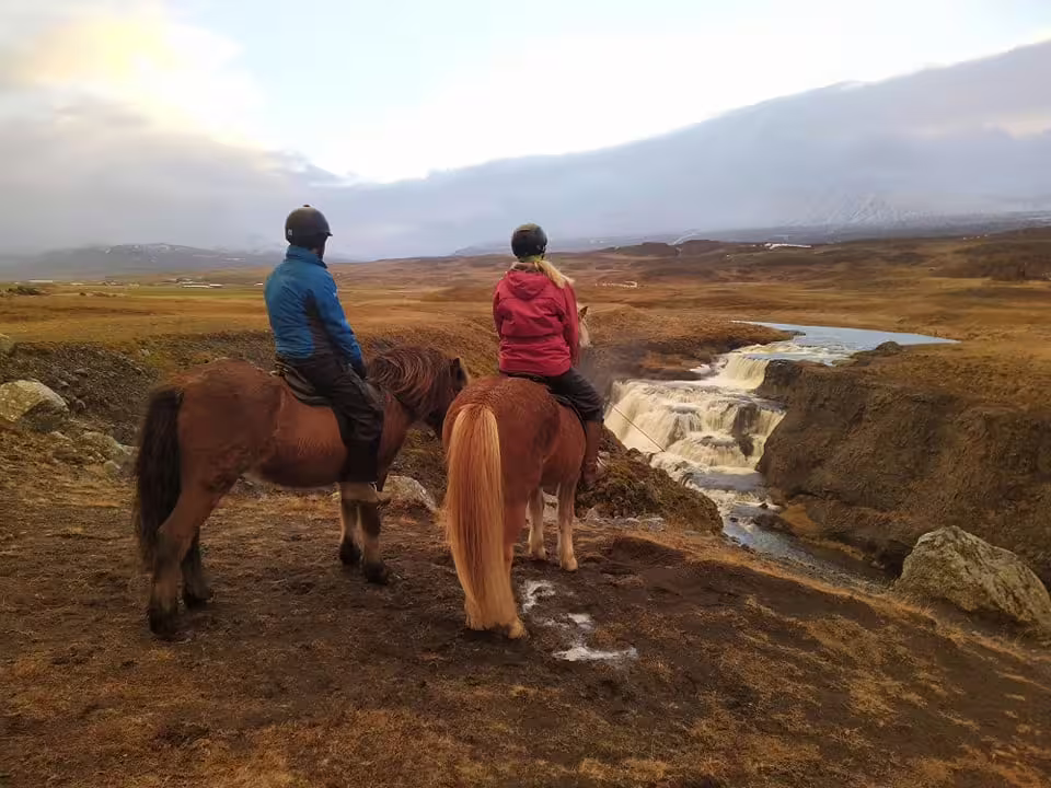 Two riders on Icelandic horses overlooking a waterfall and vast golden plains in Skagafjordur, Iceland