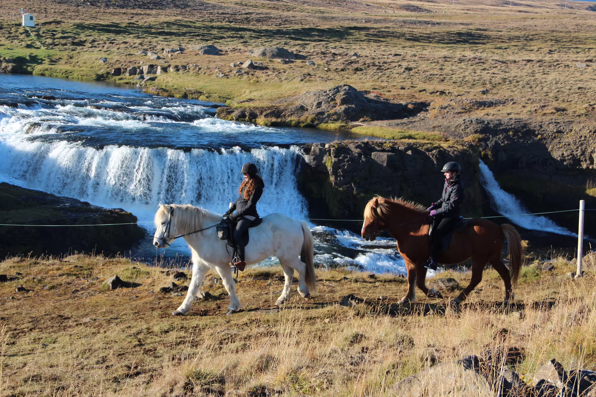 Two riders on Icelandic horses passing a cascading waterfall in Skagafjordur Iceland