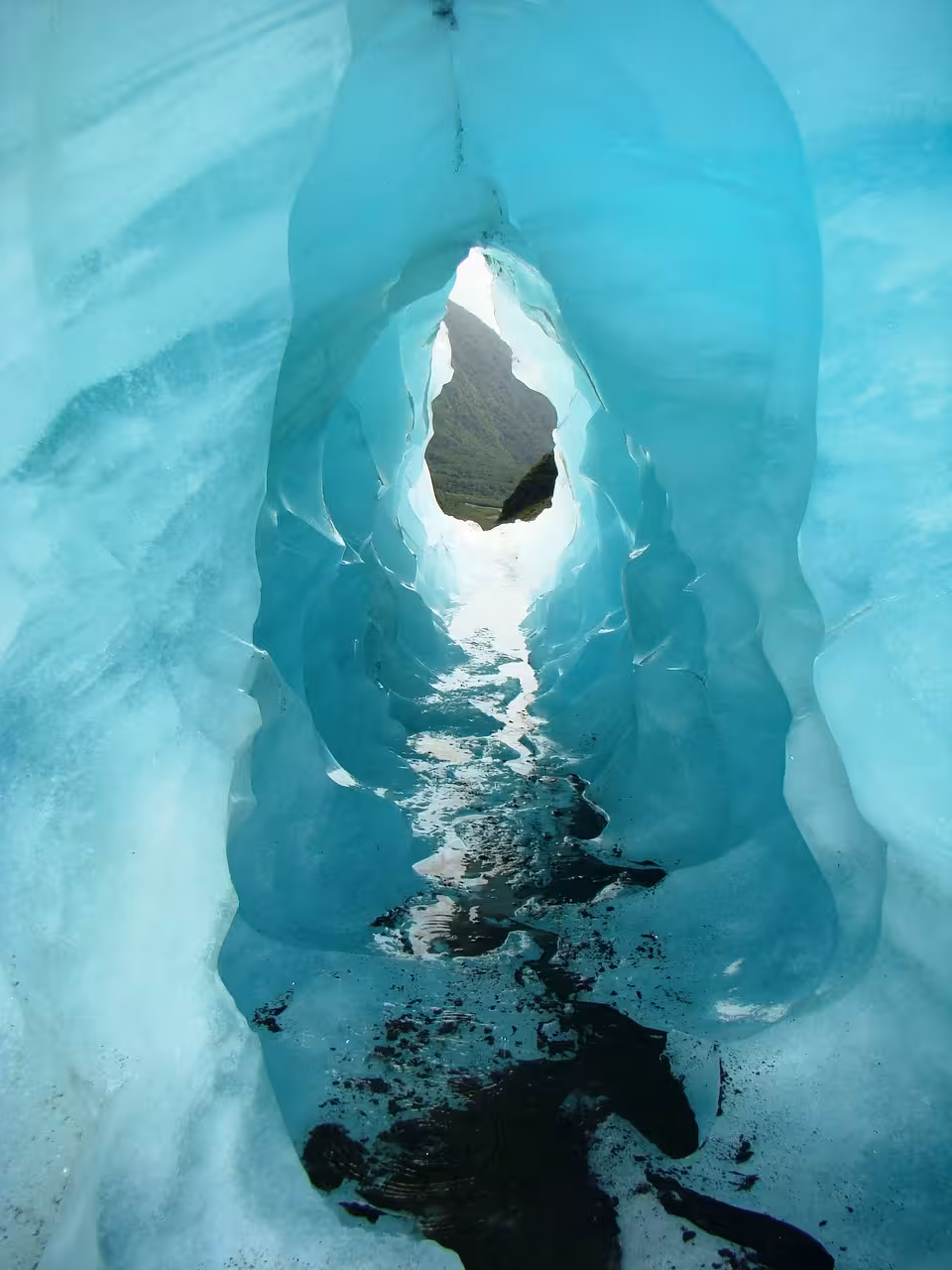 Glowing turquoise glacial ice cave tunnel with opening revealing green landscape in West Iceland