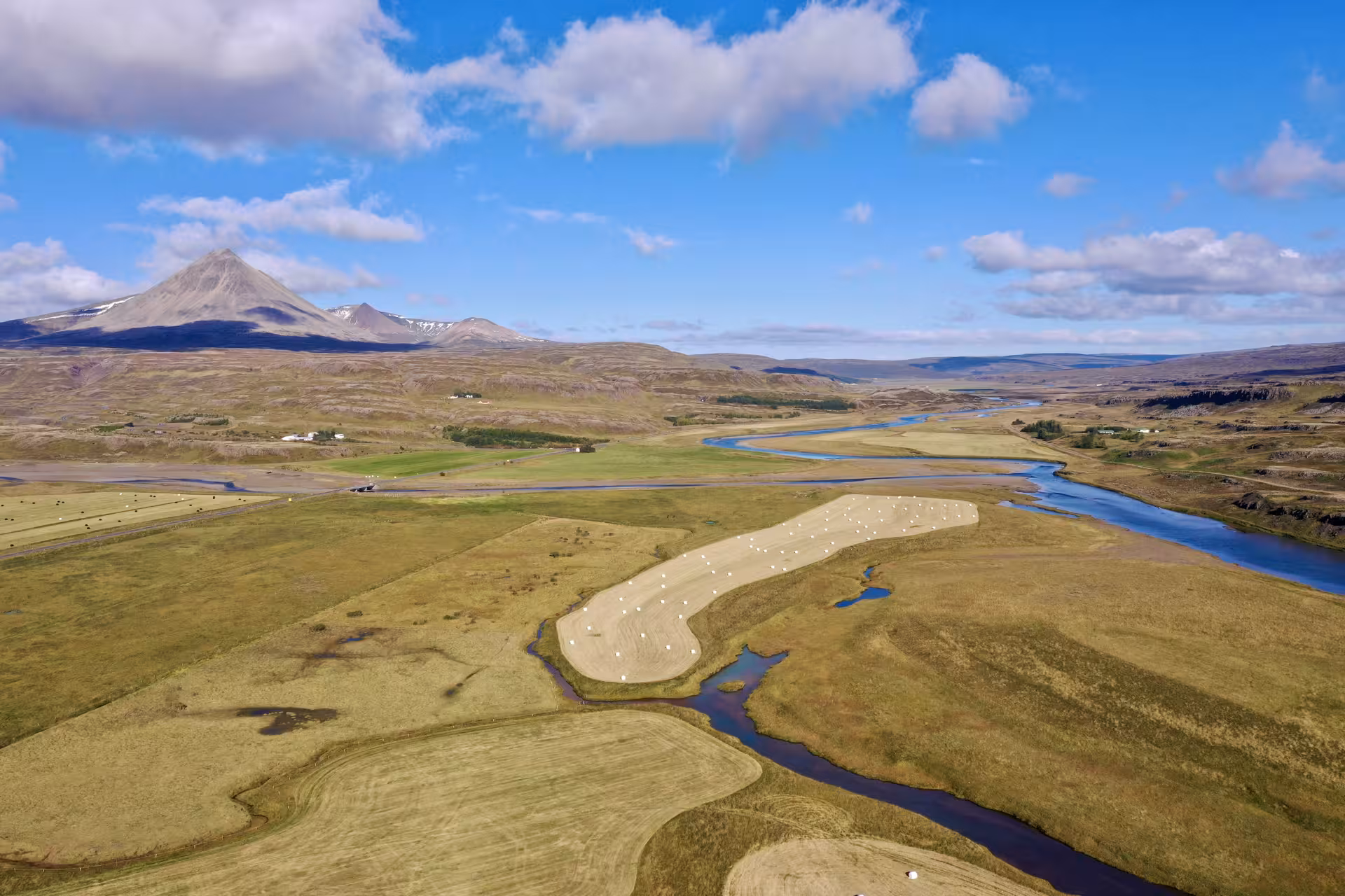 Aerial view of West Iceland river valley with hay bales, green farmland, and a volcanic mountain peak