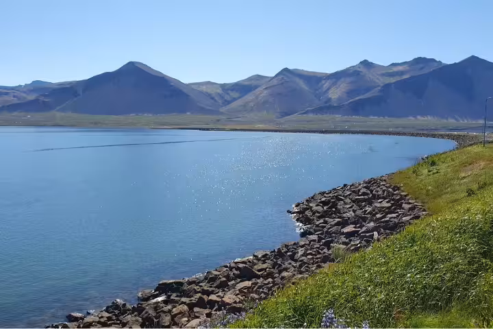 Calm blue fjord with rocky shoreline and volcanic mountains under clear sky in West Iceland