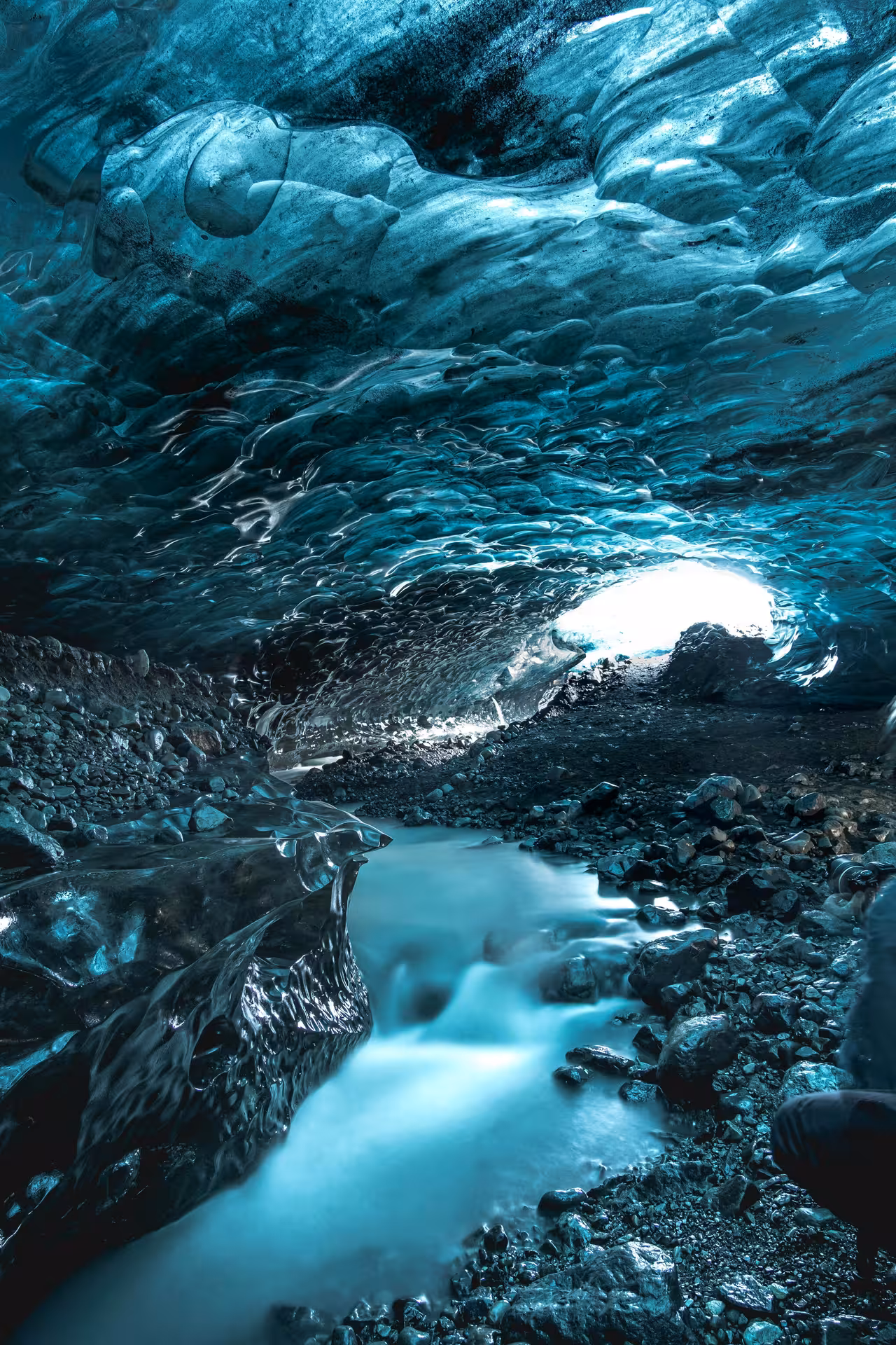 Glowing blue glacial ice cave with flowing meltwater stream and rocky floor in West Iceland