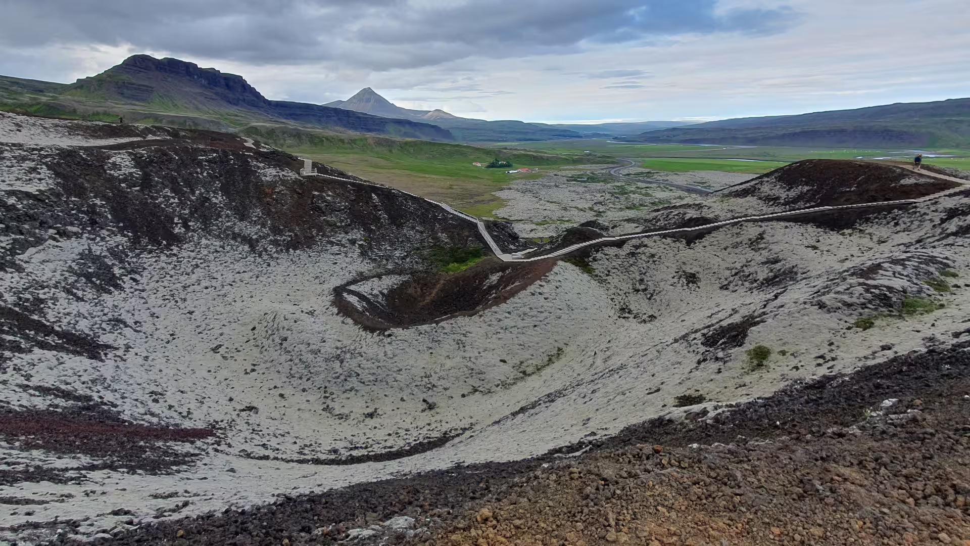 Volcanic crater with snow-dusted rim and wooden walkway, green Icelandic valley and mountain peak in background
