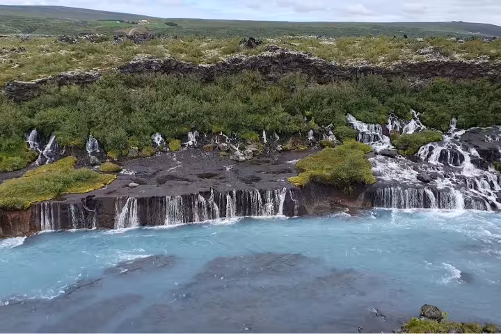 Turquoise glacial river with cascading waterfalls flowing from mossy black lava rock fields in West Iceland