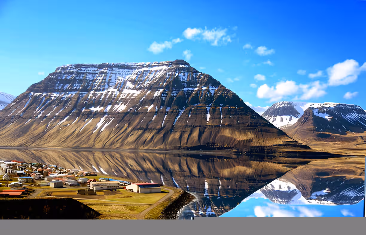 Snow-capped flat-top mountains reflecting in calm fjord waters beside a small Westfjords village, Iceland