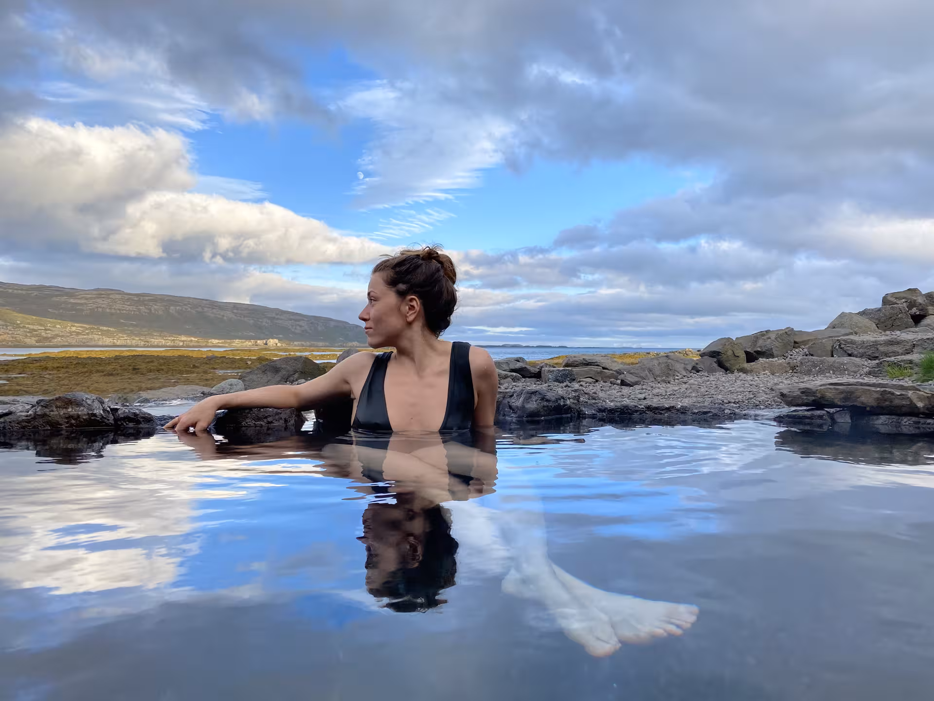 Woman relaxing in a geothermal hot spring pool with rocky Westfjords coastline and cloudy sky in Iceland