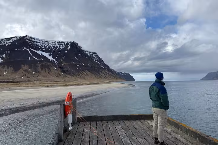 Person standing on wooden dock overlooking Westfjords coastline with snow-capped mountain and sandy beach in Iceland