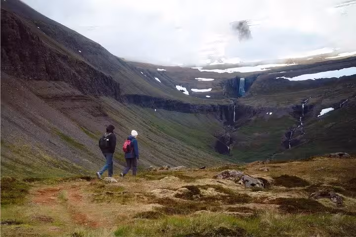 Two hikers with backpacks walking a trail toward waterfalls in the Westfjords mountains of Iceland