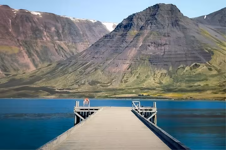 Wooden pier extending over calm blue fjord waters with dramatic layered mountains in Westfjords Iceland