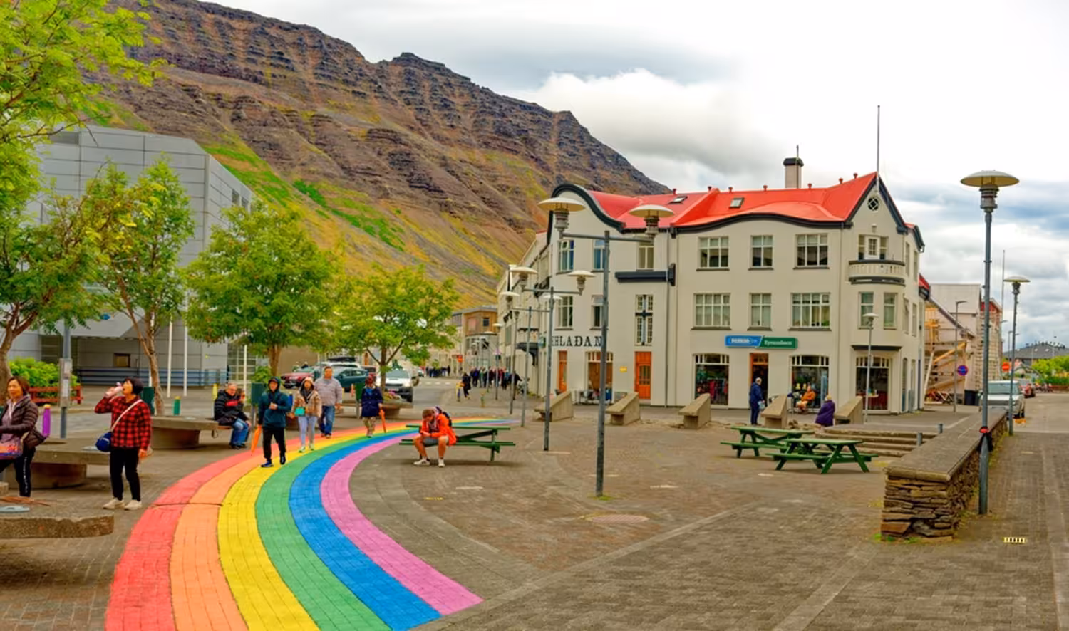 Rainbow-painted street in Westfjords town square with mountain backdrop and historic white building with red roof